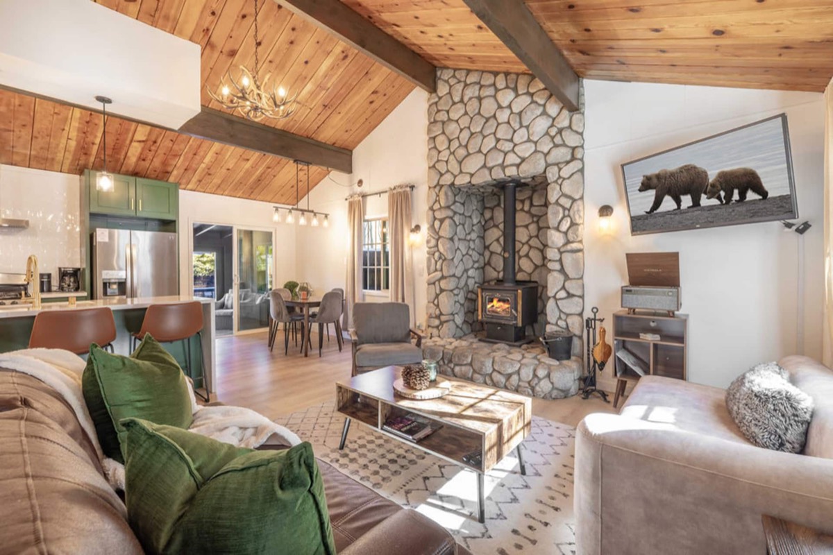 The great room: a river-rock hearth with a wood-burning stove, a vaulted knotty-pine ceiling with exposed beams, and the open kitchen beyond
