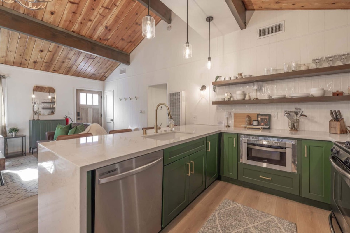 The kitchen — deep green shaker cabinets, quartz counters, open shelving and a gas range under the vaulted pine ceiling