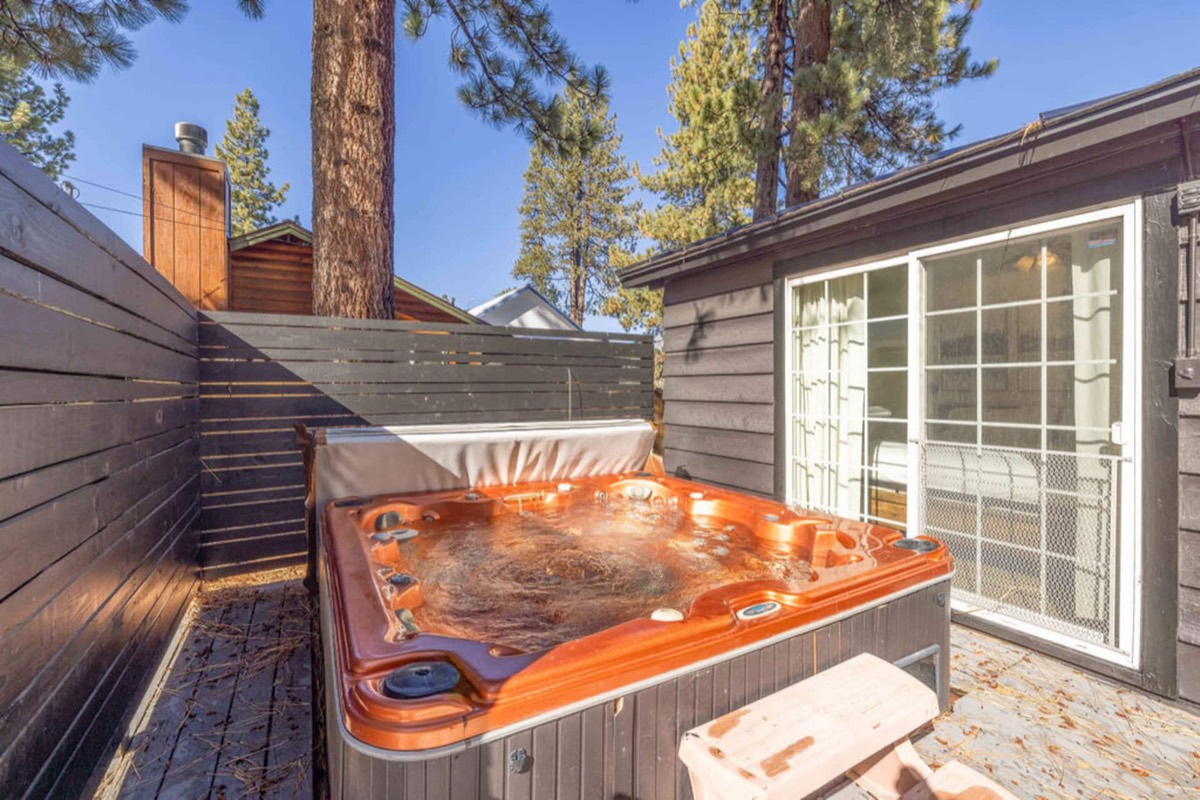 The private hot tub on the deck in a fenced yard, pines overhead and the sliding door back into the house